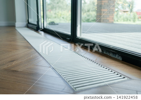 Sleek metal trench radiator in herringbone wood floor providing invisible thermal barrier near glass doors. Great image for heating system design, smart homes, and modern living spaces Sleek metal trench radiator in herringbone wood floor providing invisible thermal barrier near glass doors. Great image for heating system design, smart homes, and modern living spaces 141922458