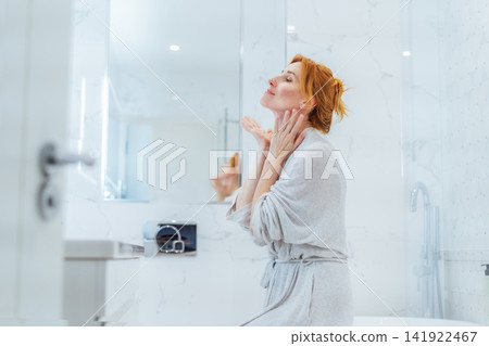 Middle aged woman doing face building yoga facial gymnastics self massage and rejuvenating exercises looking in bathroom mirror. Natural procedures for skin care. Home Beauty treatment routine Middle aged woman doing face building yoga facial gymnastics self massage and rejuvenating exercises looking in bathroom mirror. Natural procedures for skin care. Home Beauty treatment routine 141922467