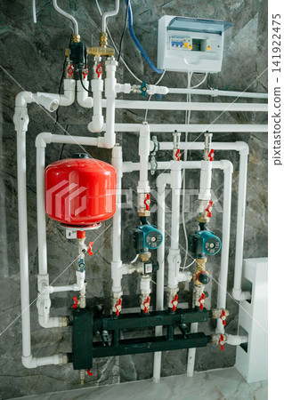 Complex piping system with pumps, pressure gauge, and red vessel installed in a utility room. Perfect stock image for home heating engineering, plumbing services, and renovation Complex piping system with pumps, pressure gauge, and red vessel installed in a utility room. Perfect stock image for home heating engineering, plumbing services, and renovation 141922475