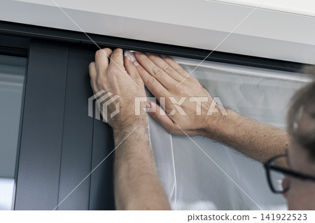 Person securing a white mosquito screen into the window frame, illustrating precise handwork in home tasks and maintenance. Person securing a white mosquito screen into the window frame, illustrating precise handwork in home tasks and maintenance. 141922523