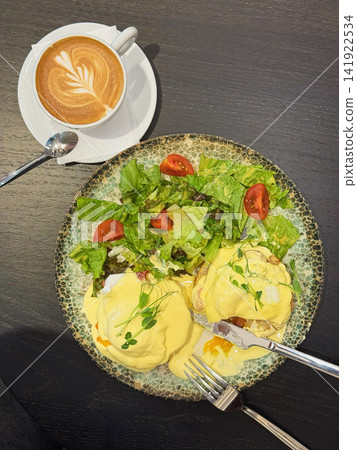 Eggs Benedict with Green Salad and Latte for Brunch. Eggs Benedict with creamy hollandaise sauce, green salad, latte served on dark cafe table. Popular meal, savory breakfast, fresh vegetables, coffee Eggs Benedict with Green Salad and Latte for Brunch. Eggs Benedict with creamy hollandaise sauce, green salad, latte served on dark cafe table. Popular meal, savory breakfast, fresh vegetables, coffee 141922534
