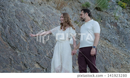 Young couple walking hand in hand along rugged hillside, woman pointing toward distance with cheerful smile while man follows beside rocky slope during outdoor travel Young couple walking hand in hand along rugged hillside, woman pointing toward distance with cheerful smile while man follows beside rocky slope during outdoor travel 141923280
