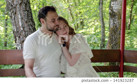 Young couple embracing on wooden bench amid sunlit forest clearing, woman resting head on man's shoulder, soft natural light, calm outdoor portrait of affection and connection Young couple embracing on wooden bench amid sunlit forest clearing, woman resting head on man's shoulder, soft natural light, calm outdoor portrait of affection and connection 141923287