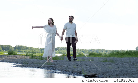 Romantic couple walking hand in hand along grassy riverbank with woman in flowing white dress gesturing beside calm water, with couple, romantic, walking, hand detail Romantic couple walking hand in hand along grassy riverbank with woman in flowing white dress gesturing beside calm water, with couple, romantic, walking, hand detail 141923302