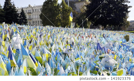 Many small blue-yellow flags with names of the dead war against russia. Memorial of the fallen soldiers, children, women in the capital of Ukraine. Concept of tragedy and misfortune. Close up Many small blue-yellow flags with names of the dead war against russia. Memorial of the fallen soldiers, children, women in the capital of Ukraine. Concept of tragedy and misfortune. Close up 141923376