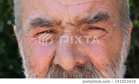 Blue eyes of elderly male blinking and looking into camera outdoor. Wrinkled face of senior human with a sad sight. Upset facial expression of pensioner. Close up Blue eyes of elderly male blinking and looking into camera outdoor. Wrinkled face of senior human with a sad sight. Upset facial expression of pensioner. Close up 141923418