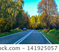 Empty Winding Asphalt Road Through Vibrant Autumn Forest Under Blue Sky in Countryside 141925057