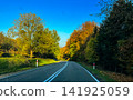 Empty Winding Asphalt Road Through Vibrant Autumn Forest Under Blue Sky in Countryside 141925059