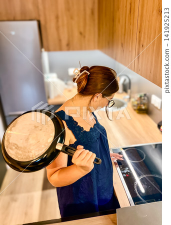 Woman Holding Frying Pan While Turning on Electric Stove Top in Modern Kitchen Woman Holding Frying Pan While Turning on Electric Stove Top in Modern Kitchen 141925213