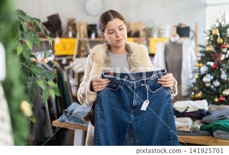 Uncertain young girl choosing jeans pantaloons in clothing store 141925701