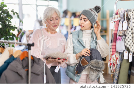 Two happy women choosing warm hat for stylish autumn look in clothing boutique 141925725