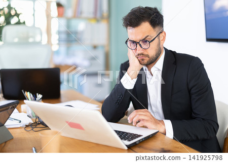 young man in glasses sits at a table and looks at a laptop screen young man in glasses sits at a table and looks at a laptop screen 141925798