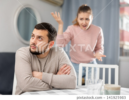 Upset guy sitting at table with frustrated girlfriend in background Upset guy sitting at table with frustrated girlfriend in background 141925983