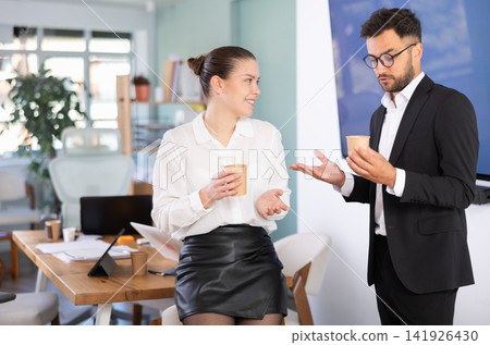Man and a woman work in an office, they stand near a table and drink coffee Man and a woman work in an office, they stand near a table and drink coffee 141926430