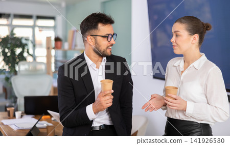 Man and a woman work in an office, they stand near a table and drink coffee Man and a woman work in an office, they stand near a table and drink coffee 141926580