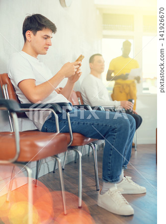 Guy teenager waiting in line at reception with phone in his hand Guy teenager waiting in line at reception with phone in his hand 141926670