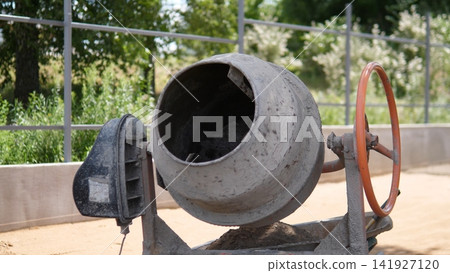 Dusty concrete mixer drum stands outdoors at construction site, showing worn metal housing, orange hand wheel, and dark opening, with soft green trees and bright daylight behind Dusty concrete mixer drum stands outdoors at construction site, showing worn metal housing, orange hand wheel, and dark opening, with soft green trees and bright daylight behind 141927120