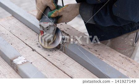 Close view of gloved hands guiding grinder across steel bar on weathered wooden boards, creating sparks for fabrication, repair, welding prep, maintenance, and workshop construction Close view of gloved hands guiding grinder across steel bar on weathered wooden boards, creating sparks for fabrication, repair, welding prep, maintenance, and workshop construction 141927149