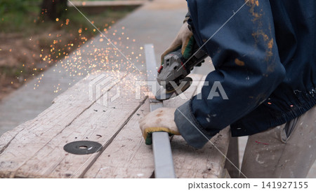 Close view of worker shaping metal pipe with handheld grinder on weathered wooden bench outdoors, bright orange sparks flying through air, worn gloves, practical workwear, and focused manual task Close view of worker shaping metal pipe with handheld grinder on weathered wooden bench outdoors, bright orange sparks flying through air, worn gloves, practical workwear, and focused manual task 141927155