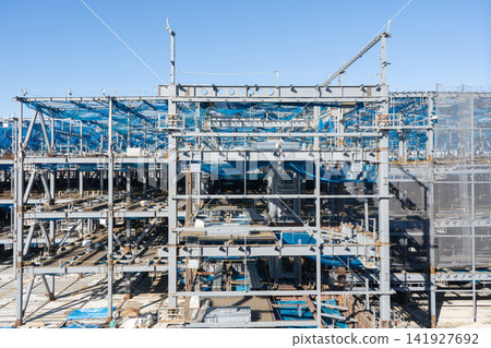 A large-scale construction site with a multi-story steel-frame building and blue safety nets installed. 141927692