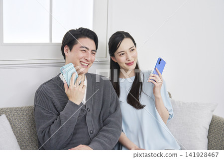 A portrait of a couple in their living room, holding a smartphone and looking directly at the camera. A portrait of a couple in their living room, holding a smartphone and looking directly at the camera. 141928467
