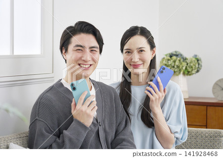 A portrait of a couple in their living room, holding a smartphone and looking directly at the camera. A portrait of a couple in their living room, holding a smartphone and looking directly at the camera. 141928468