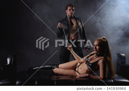 Provocative couple in black leather playing dominance game with chain in smoky studio Provocative couple in black leather playing dominance game with chain in smoky studio 141930968