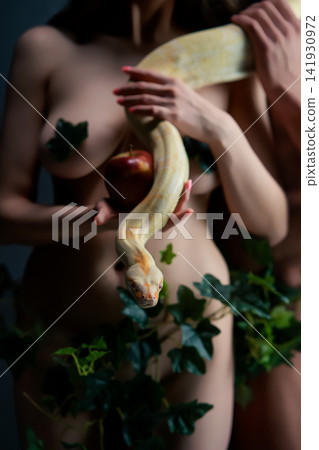 Nude woman holding apple with yellow snake coiled around her body surrounded by green ivy leaves 141930972