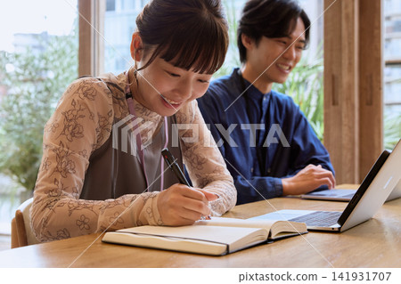 一對年輕的商務人士,一男一女,微笑著在咖啡館裡用筆記本寫作。 一對年輕的商務人士,一男一女,微笑著在咖啡館裡用筆記本寫作。 141931707