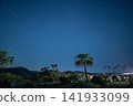 A night sky with shooting stars, a single palm tree, and the lights of a campsite. 141933099