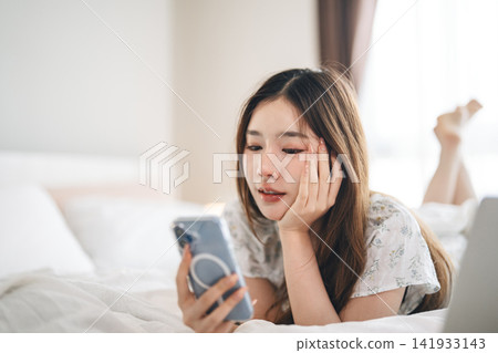 Young adult asian woman wearing pajamas in white bedroom on morning scrolling smartphone resting Young adult asian woman wearing pajamas in white bedroom on morning scrolling smartphone resting 141933143