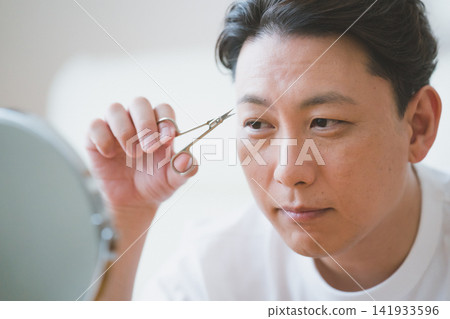 A 40-year-old man trims his eyebrows with eyebrow scissors while looking in the mirror. A 40-year-old man trims his eyebrows with eyebrow scissors while looking in the mirror. 141933596