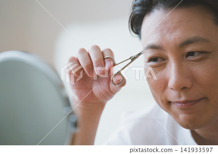 A smiling 40-year-old man grooms his eyebrows with eyebrow scissors: Men's eyebrow care. A smiling 40-year-old man grooms his eyebrows with eyebrow scissors: Men's eyebrow care. 141933597