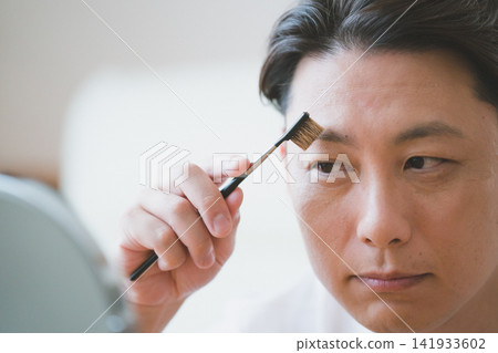 一位四十多歲的男子正在對著鏡子用眉刷修眉。 一位四十多歲的男子正在對著鏡子用眉刷修眉。 141933602