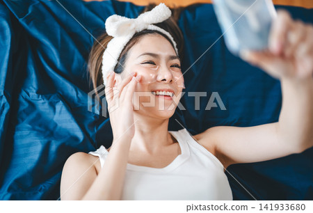 Beautiful smiling young asian woman, relax lying in bed with eye sheet mask enjoy skincare Beautiful smiling young asian woman, relax lying in bed with eye sheet mask enjoy skincare 141933680
