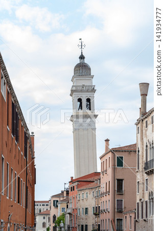 Tall white campanile rising above narrow Venetian street under cloudy sky 141934777