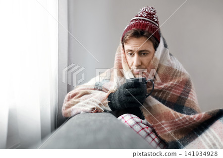 freezing man wrapped in a blanket holds a hot drink at home. He suffers from a cold snap, frost, and weak heating due to the energy crisis and heating costs. freezing man wrapped in a blanket holds a hot drink at home. He suffers from a cold snap, frost, and weak heating due to the energy crisis and heating costs. 141934928