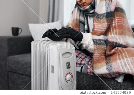 freezing man in a blanket uses a heater at home during winter, illustrating the energy crisis, frost, and rising heating costs. freezing man in a blanket uses a heater at home during winter, illustrating the energy crisis, frost, and rising heating costs. 141934929
