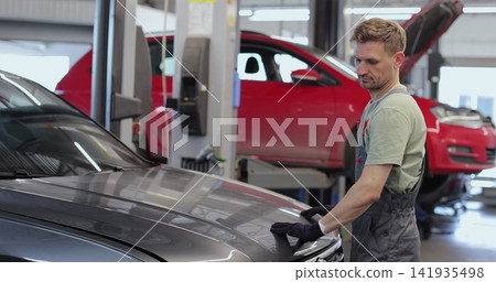 Professional car mechanic is working on a vehicle in a service. Happy repairman is closing the hood. Man Looks at a Camera and Smiles. Professional car mechanic is working on a vehicle in a service. Happy repairman is closing the hood. Man Looks at a Camera and Smiles. 141935498
