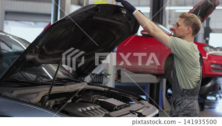 Professional car mechanic is working on a vehicle in a service. Happy repairman is closing the hood. Man Looks at a Camera and Smiles. Professional car mechanic is working on a vehicle in a service. Happy repairman is closing the hood. Man Looks at a Camera and Smiles. 141935500