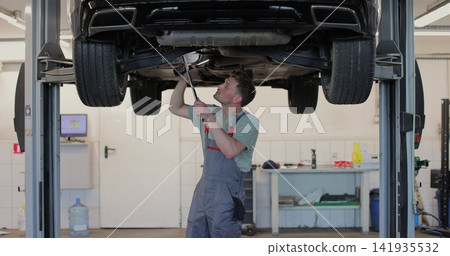 Car mechanic is working under a vehicle on a lift in service. Repairman is using ratchet. Car mechanic is working under a vehicle on a lift in service. Repairman is using ratchet. 141935532