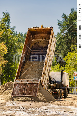 A dump truck tilts its bed to release a load of sand at a construction site. The area features trees and a road sign, with clear visibility A dump truck tilts its bed to release a load of sand at a construction site. The area features trees and a road sign, with clear visibility 141936406