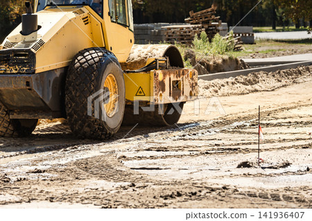 A large yellow road roller is on a construction site with fresh dirt and marked spots. Nearby, construction materials are stacked for future use A large yellow road roller is on a construction site with fresh dirt and marked spots. Nearby, construction materials are stacked for future use 141936407