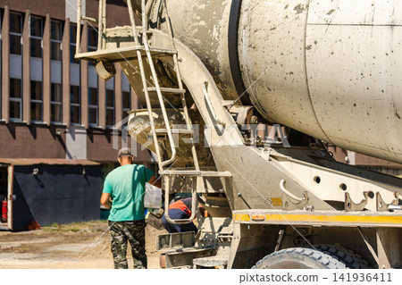 A concrete mixer truck stands at a construction site. Workers are preparing concrete for use in ongoing building work. Several materials are scattered around A concrete mixer truck stands at a construction site. Workers are preparing concrete for use in ongoing building work. Several materials are scattered around 141936411