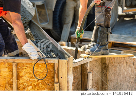 Workers are using a hose to pour concrete into a wooden form. One worker stands on the form while another supports the hose Workers are using a hose to pour concrete into a wooden form. One worker stands on the form while another supports the hose 141936412