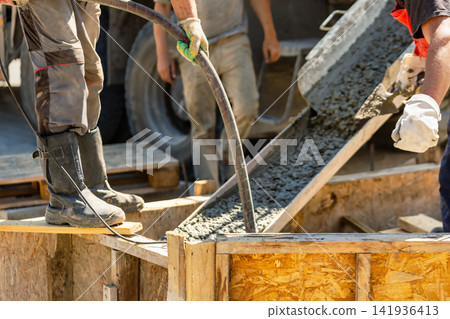 Two workers use hoses to pour concrete into a wooden structure at a construction site during the afternoon. Heavy equipment is in the background Two workers use hoses to pour concrete into a wooden structure at a construction site during the afternoon. Heavy equipment is in the background 141936413