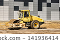 A yellow roller compacts dirt on a construction site next to a building with a gray and black facade. Workers are present in the background 141936414