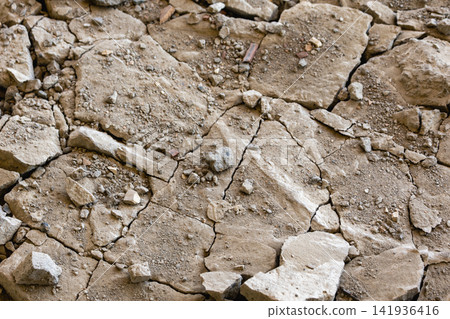 Cracked soil shows large broken pieces and small stones scattered across the ground in a dry area. The earth appears arid and tough Cracked soil shows large broken pieces and small stones scattered across the ground in a dry area. The earth appears arid and tough 141936416