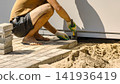 A person works on laying tiles on a ground surface using a trowel. The area has sand and building materials close to a white wall during the day 141936419