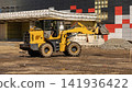 A construction vehicle is working on a site where workers are arranging materials. The area is full of dirt, with piles of gravel and other supplies present 141936422